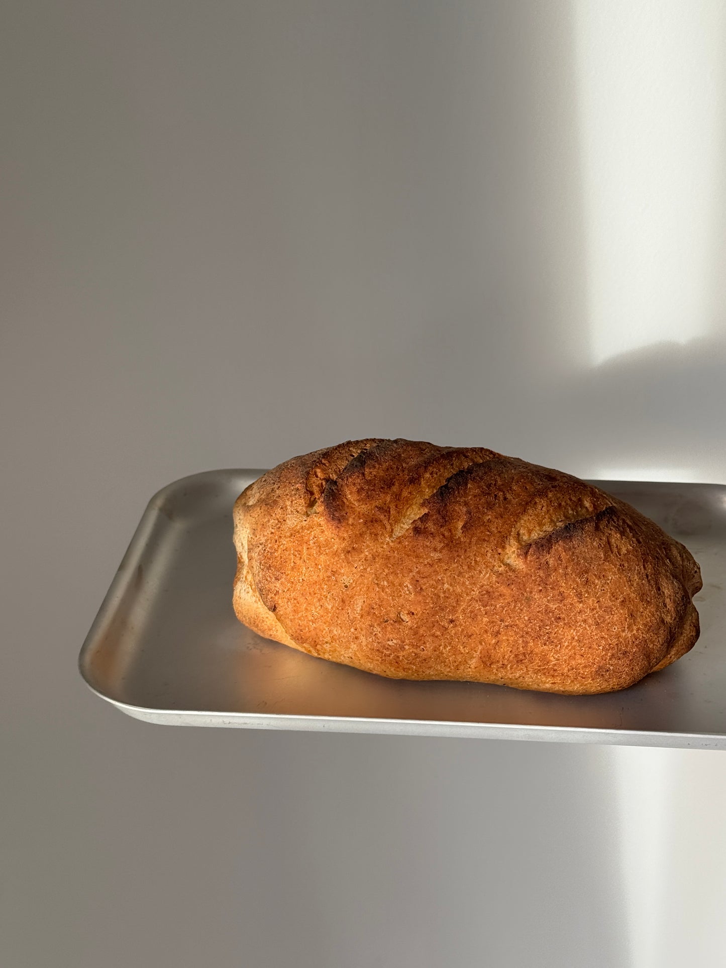 A single rustic gluten free bread loaf with a golden-brown crust sits on a silver metal tray against a plain light gray background.