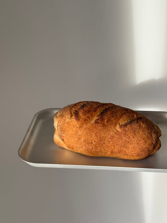 A single rustic gluten free bread loaf with a golden-brown crust sits on a silver metal tray against a plain light gray background.