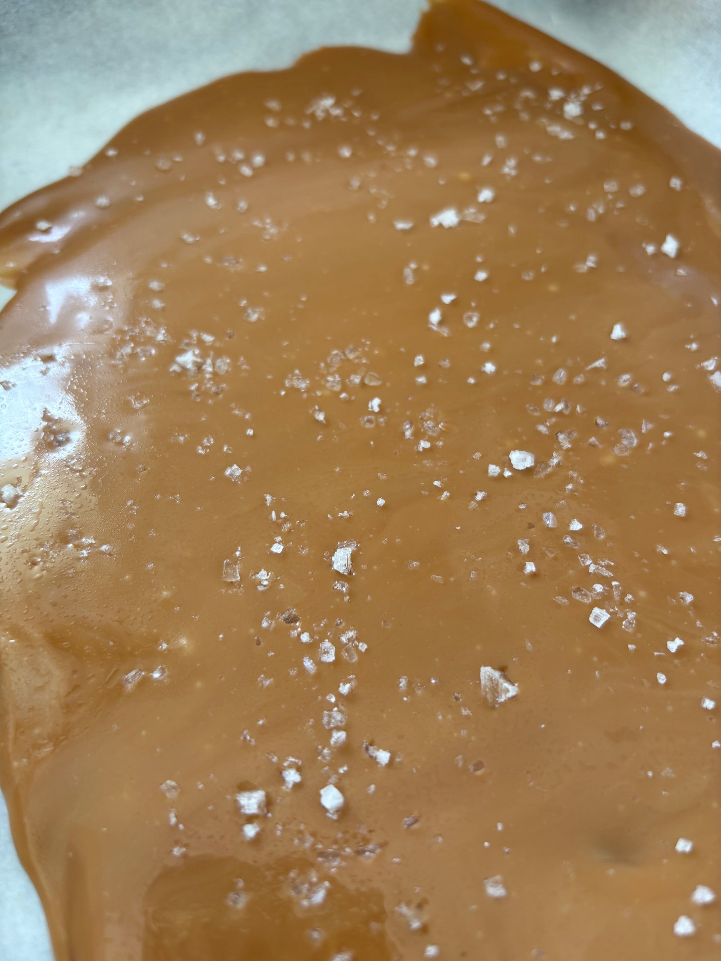 A close-up of a glossy brown caramel or surface sprinkled with coarse white sea salt, photographed from above.
